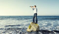 Concept of looking for something. Man in jeans and shirt stands on a rock in the middle of the sea. He is holding a telescope and looking at something.