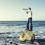 Concept of looking for something. Man in jeans and shirt stands on a rock in the middle of the sea. He is holding a telescope and looking at something.