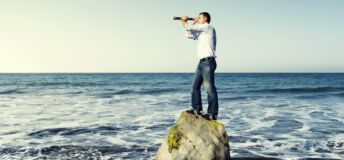 Concept of looking for something. Man in jeans and shirt stands on a rock in the middle of the sea. He is holding a telescope and looking at something.