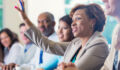 Mid adult African American businesswoman is smiling and raising her hand. She is answering a question during a business conference or job training seminar. Professional woman is wearing