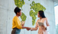 Two diverse businesswomen shaking hands, celebrating a successful business deal in a modern office featuring a unique moss wall map, embodying collaboration and mutual respect