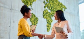 Two diverse businesswomen shaking hands, celebrating a successful business deal in a modern office featuring a unique moss wall map, embodying collaboration and mutual respect