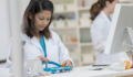 Focus is on a serious young pharmacist as she looks down and concentrates on counting prescription medication. There are prescription bottles and a computer on the table and another pharmacist and shelves in the background.