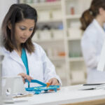 Focus is on a serious young pharmacist as she looks down and concentrates on counting prescription medication. There are prescription bottles and a computer on the table and another pharmacist and shelves in the background.
