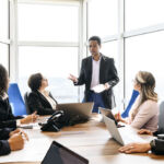 Group of business professionals sitting around table with laptops and digital tablets, listening to male manager in his 30s, serious, brainstorming, collaboration