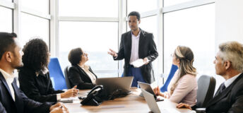 Group of business professionals sitting around table with laptops and digital tablets, listening to male manager in his 30s, serious, brainstorming, collaboration