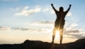 Man with backpack putting his hands up and standing on cliff at sunset time, sun rays on background with clouds, warm tone and almost in silhouette photo
