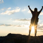Man with backpack putting his hands up and standing on cliff at sunset time, sun rays on background with clouds, warm tone and almost in silhouette photo