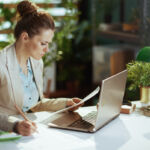 modern woman worker in a light business suit in modern green office with documents and laptop.