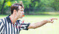 Handsome mid adult Caucasian referee points as he blows his whistle. He is calling a penalty or foul during sporting event. He has brown hair and is wearing a black and white striped referee uniform. He is standing on the playing field. He has an intense expression on his face.