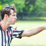 Handsome mid adult Caucasian referee points as he blows his whistle. He is calling a penalty or foul during sporting event. He has brown hair and is wearing a black and white striped referee uniform. He is standing on the playing field. He has an intense expression on his face.