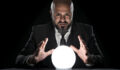 A fortune teller sitting at a desk gesturing at a glowing crystal ball. The bald man with the long beard wearing an elegant black suit, a white button down shirt and a tie. He is staring concentrated at the camera.