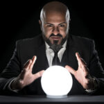 Wanna bet? The risks of predictive markets A fortune teller sitting at a desk gesturing at a glowing crystal ball. The bald man with the long beard wearing an elegant black suit, a white button down shirt and a tie. He is staring concentrated at the camera.