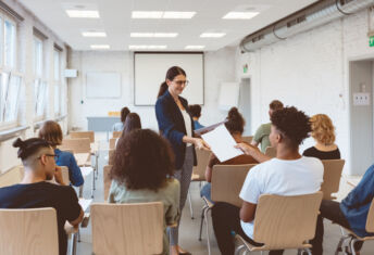 Female tutor giving test papers to male student in classroom. Professor is teaching at university. She is wearing smart casuals.