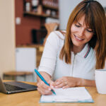 Shot of an adorable smiling businesswoman using laptop and making notes on a clipboard at home.