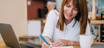 Shot of an adorable smiling businesswoman using laptop and making notes on a clipboard at home.