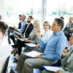 Large group of business people attending a seminar in a board room. Focus is on mid adult businessman in blue shirt.