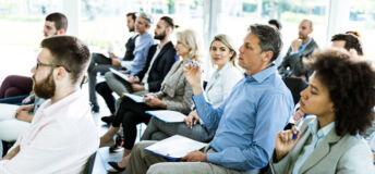 Large group of business people attending a seminar in a board room. Focus is on mid adult businessman in blue shirt.