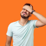 Facepalm. Portrait of desperate brunette man with beard in white t-shirt covering face with hand, feeling sorry and blaming himself for the mistake. indoor studio shot isolated on orange background