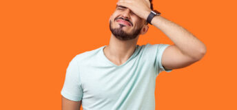 Facepalm. Portrait of desperate brunette man with beard in white t-shirt covering face with hand, feeling sorry and blaming himself for the mistake. indoor studio shot isolated on orange background