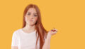 A young woman with long red hair expresses uncertainty, twirling her hair while standing in front of a vibrant yellow backdrop in a bright indoor setting.