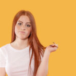 A young woman with long red hair expresses uncertainty, twirling her hair while standing in front of a vibrant yellow backdrop in a bright indoor setting.