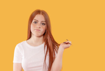A young woman with long red hair expresses uncertainty, twirling her hair while standing in front of a vibrant yellow backdrop in a bright indoor setting.