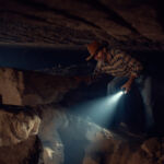 Adult man with hat and flashlight exploring a deep, dark underground cavern
