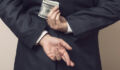 Businessman in a suit holding his hands behind his back with stack of dollar banknotes in one and fingers crossed on another one