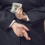 Businessman in a suit holding his hands behind his back with stack of dollar banknotes in one and fingers crossed on another one