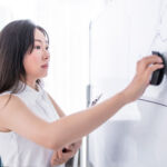 SIFMA language for revising B&R rule This indoor bright office scene shows a young adult Asian woman with long dark hair wearing a white sleeveless shirt, using a black eraser to clean written marks off a large whiteboard. She has a focused, concentrated facial expression while working in the tidy, naturally lit workspace near a window with sheer curtains.