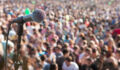 A microphone on the left size of the frame and the entire background is a blurry, out of focus crowd waiting for the next performer or speaker at an outdoor music festival.