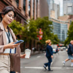 Beautiful young Japanese Asian businesswoman using her smart phone and drinking coffee to go while crossing the street in Sydney in Australia.