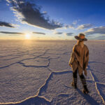 Female tourist, wearing a traditional hat and poncho, admires the sunset at Salar de Uyuni, Altiplano, Bolivia. Salar de Uyuni (or Salar de Tunupa) is the world's largest salt flat at 10,582 square kilometers (4,086 sq mi). It is located in the Potosi and Oruro departments in southwest Bolivia, near the crest of the Andes, and is elevated 3,656 meters above the mean sea level. The Salar was formed as a result of transformations between several prehistoric lakes.