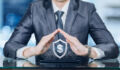A businessman is sitting at a glass table and holding his hands in protective position under a shield with currency unit inside with networking connections standing on a gadget at blurred background.