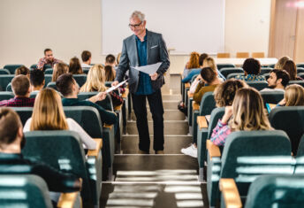 Happy senior professor talking to his students while giving them test results in lecture hall.