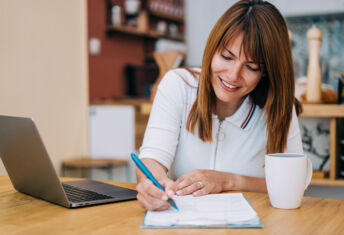 Shot of an adorable smiling businesswoman using laptop and making notes on a clipboard at home.